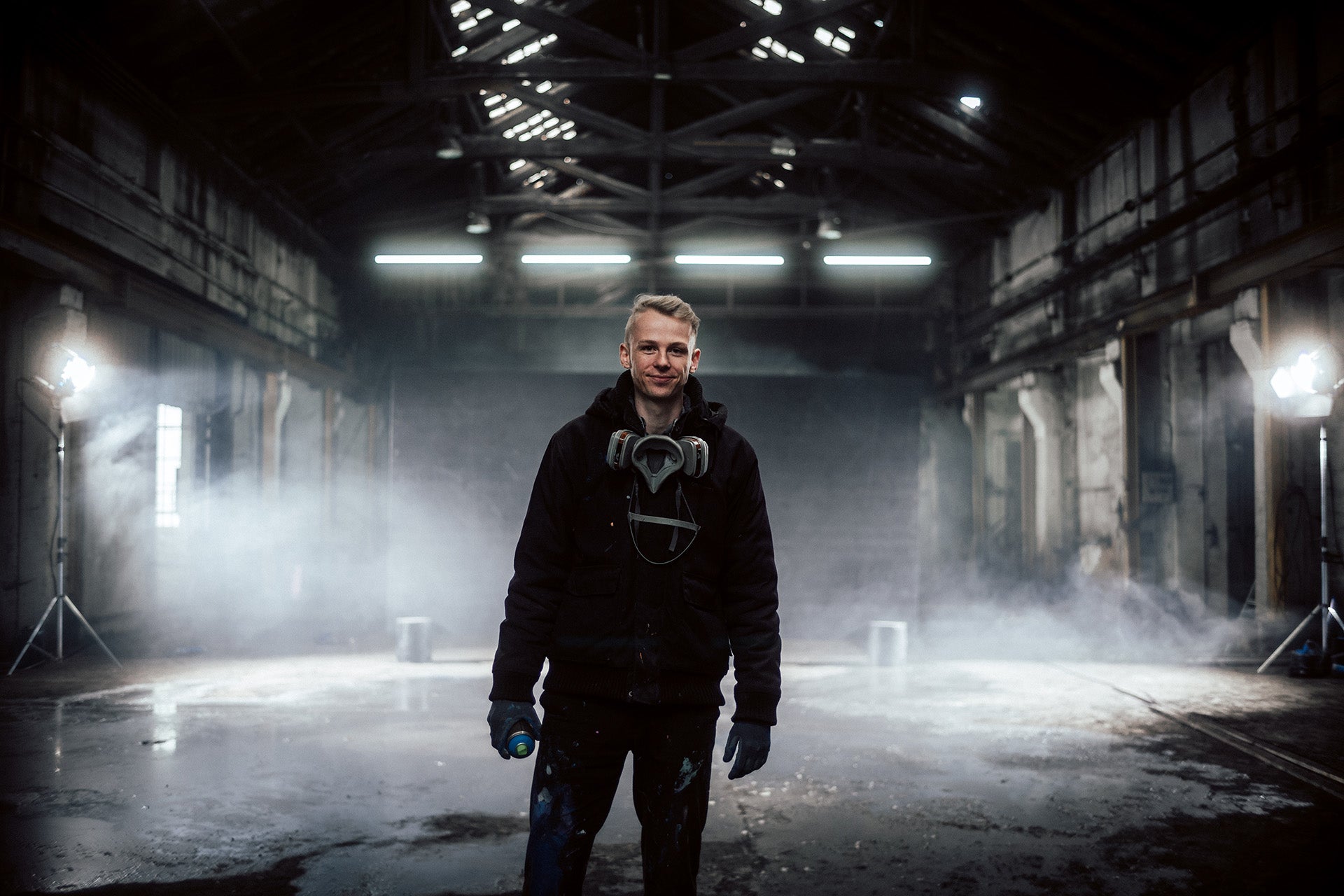Das Bild zeigt einen Künstler in einer verlassenen, stimmungsvoll beleuchteten Industriehalle. Er trägt dunkle Kleidung, eine Atemschutzmaske um den Hals und hält eine Sprühdose in der Hand – ein klarer Hinweis auf seine Arbeit als Street Artist oder Graffitikünstler. Die Szenerie ist von Nebel durchzogen, was dem Moment eine besondere Intensität und Ruhe verleiht.
