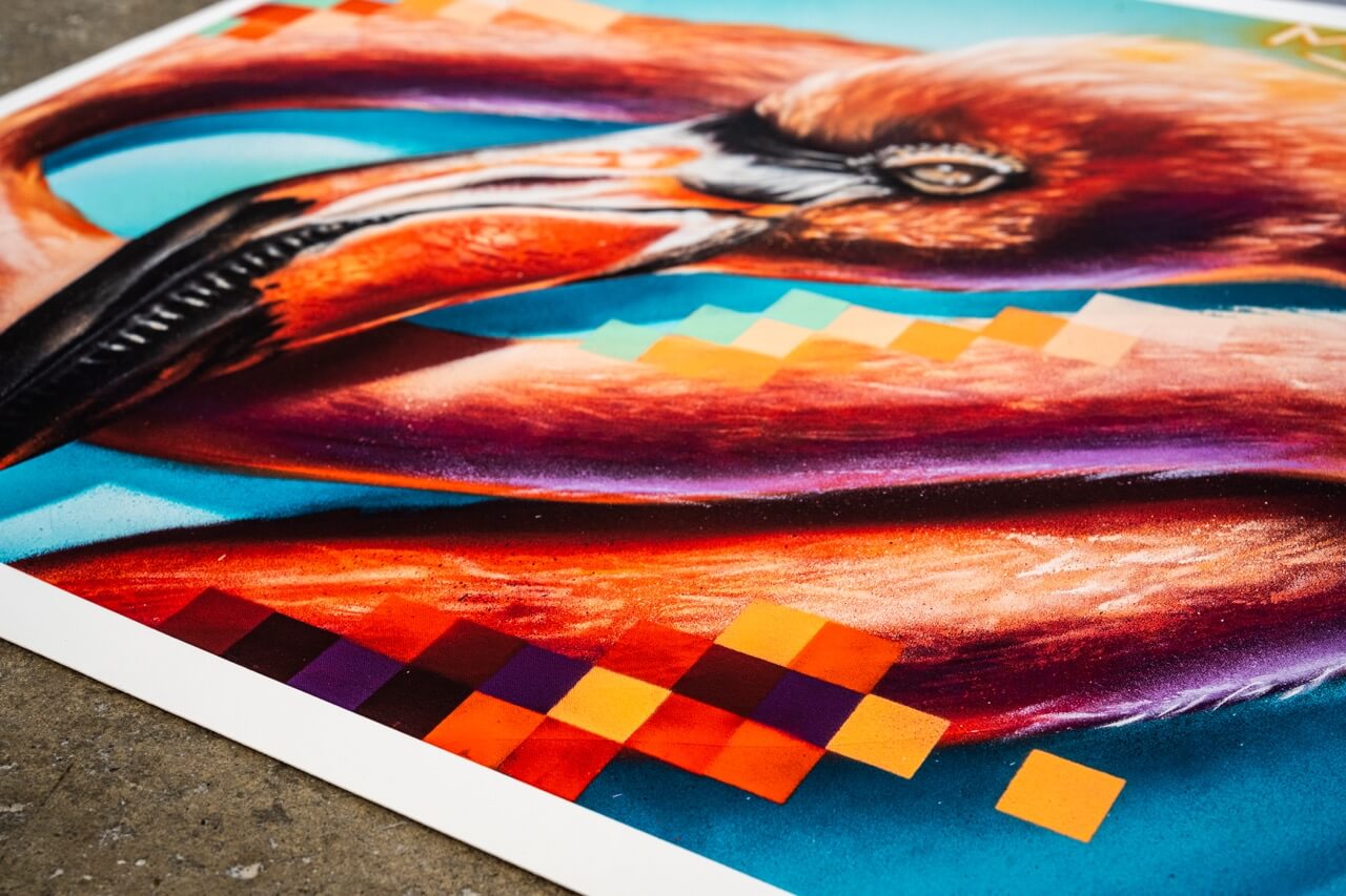 Nahaufnahme eines hochwertigen Kunstdrucks von Stone Graffiti. Das Motiv zeigt einen detailliert gemalten Flamingo mit realistischem Schnabel und fein ausgearbeiteten Federn in leuchtenden Orangetönen. Der Hintergrund enthält pixelartige Farbakzente in Blau, Orange und Lila, die eine moderne, digitale Ästhetik erzeugen. Der Druck liegt auf einem Betonboden, wodurch die künstlerische Street-Art-Atmosphäre unterstrichen wird.