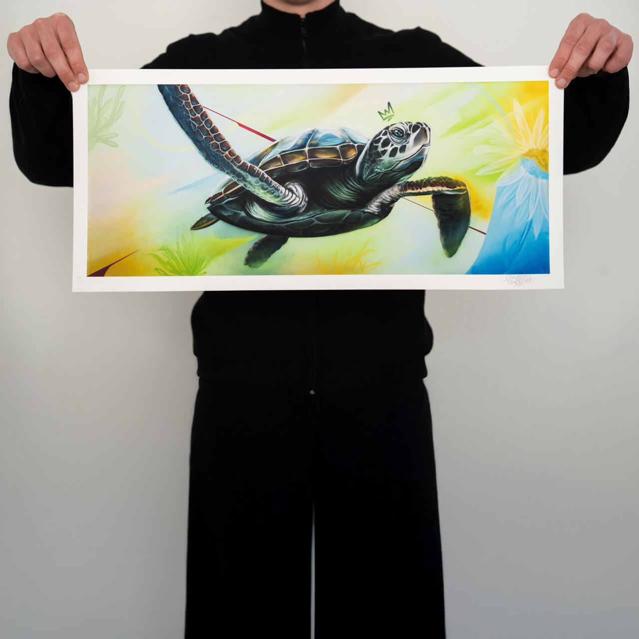 Ein hochwertiger Kunstdruck von Stone Graffiti, gehalten von einer Person in schwarzer Kleidung. Das Motiv zeigt eine beeindruckend detaillierte Meeresschildkröte, die durch eine farbenfrohe, abstrakte Unterwasserwelt schwimmt. Feine Schattierungen und Farbübergänge verleihen dem Kunstwerk Tiefe und Dynamik. Die moderne Interpretation kombiniert realistische Elemente mit grafischen Akzenten, darunter eine kleine Krone über dem Kopf der Schildkröte. Der weiße Rand des Drucks setzt das Motiv perfekt in Szene.