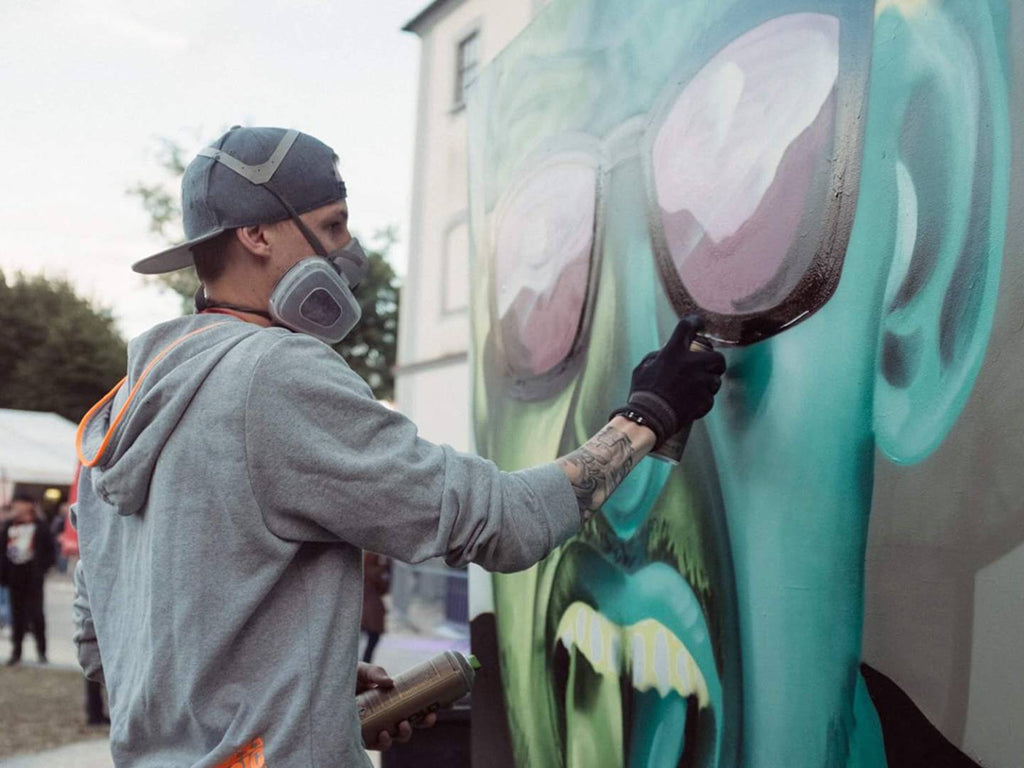 Das Bild zeigt einen Graffitikünstler bei der Arbeit an einem großflächigen Wandgemälde. Mit Atemschutzmaske, Cap und Handschuhen ausgerüstet, trägt er konzentriert Farbe auf die übergroße Sonnenbrille einer surrealen Figur auf. Die Szene wirkt urban und kreativ, im Hintergrund stehen neugierige Zuschauer.