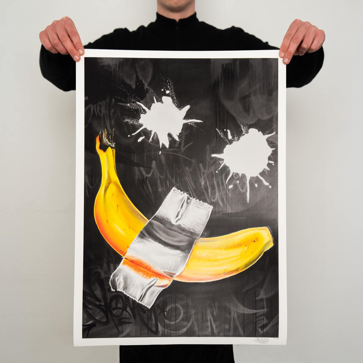 Eine Person hält den limitierten Banane Graffiti Kunstdruck mit Klebeband und weißen Farbspritzern