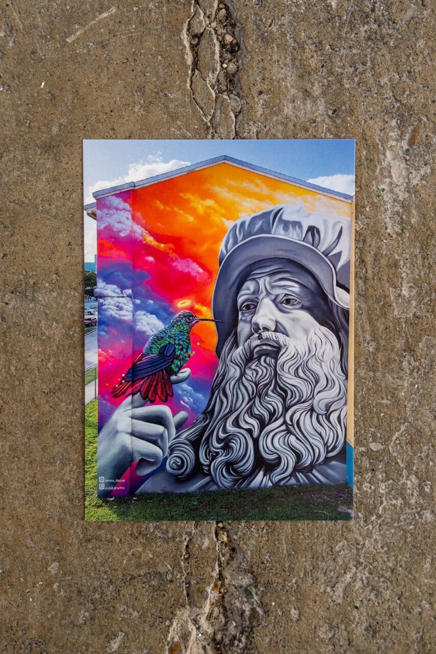 Ein farbgewaltiges Statement an der Hauswand: Diese Postkarte zeigt ein monumentales Mural, das Klassik und Urban Art vereint. Die Statue im Stil eines Renaissance-Meisters wird von einem leuchtenden Kolibri besucht – Symbol für Inspiration, Leichtigkeit und Wandel. Ein echter Hingucker für Kunstliebhaber*innen, die Gegensätze feiern.