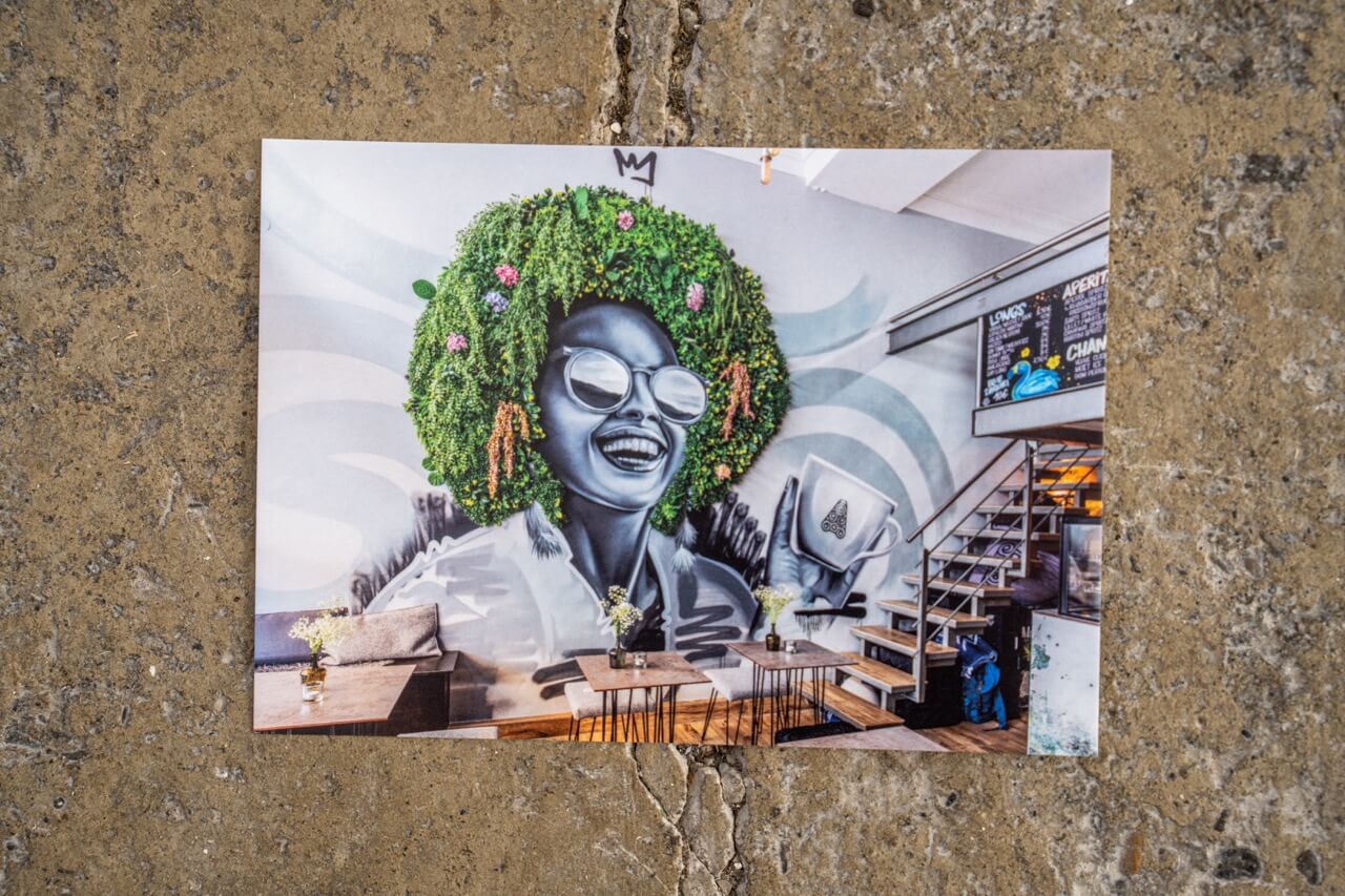 Diese Postkarte bringt gute Laune an jede Wand: Ein überdimensionales Wandbild in einem Café, das Lebensfreude pur ausstrahlt. Die afro-grüne Haarpracht aus echten Pflanzen, das strahlende Lachen und die detailverliebte Umsetzung machen das Artwork zu einem echten Hingucker – urban, originell und voller Energie.