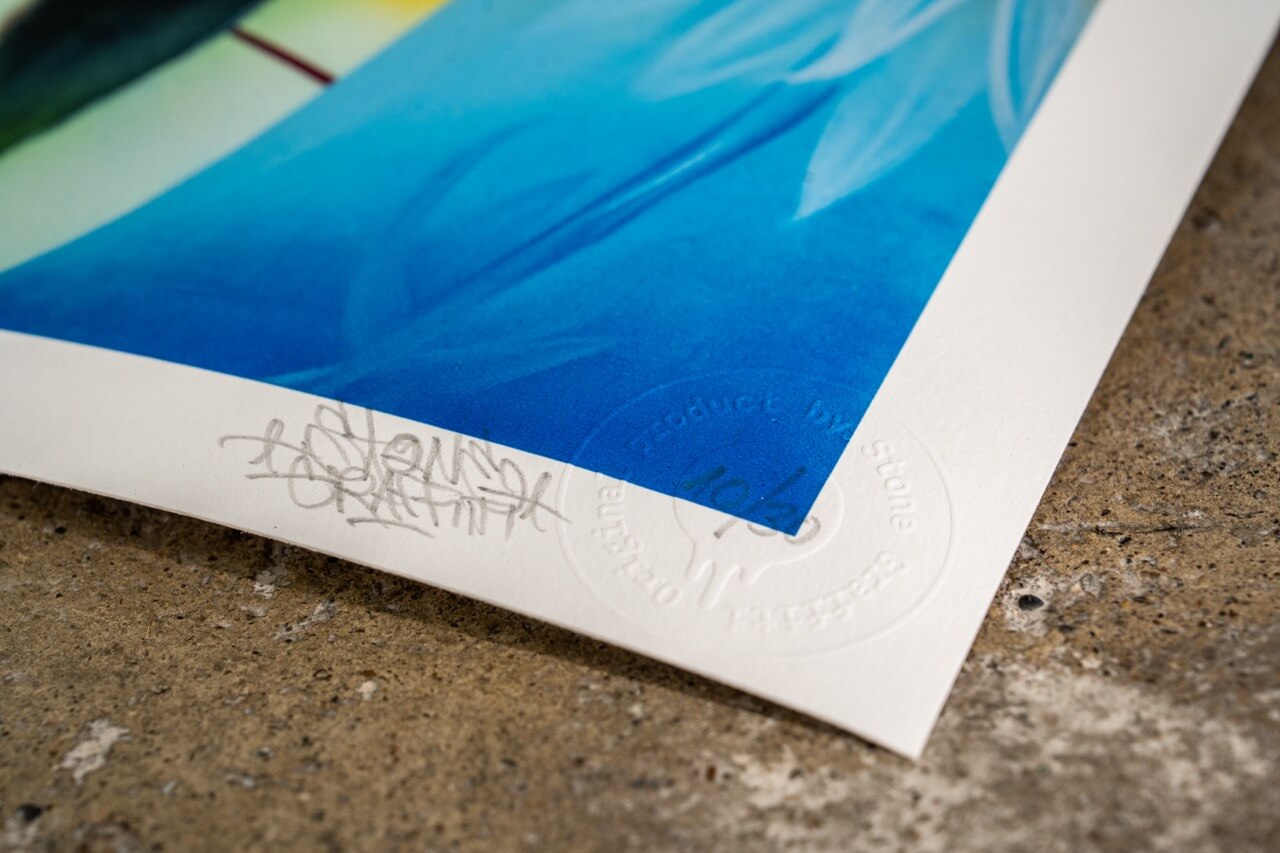 Detailaufnahme eines signierten Kunstdrucks von Stone Graffiti mit einem kräftigen blauen Farbübergang. Der Druck zeigt die handschriftliche Signatur des Künstlers sowie eine geprägte Echtheitsprägung im unteren Rand. Die Textur des hochwertigen Papiers ist erkennbar, und das Bild liegt auf einem Betonboden, der einen interessanten Kontrast zur glatten Oberfläche des Drucks bildet.