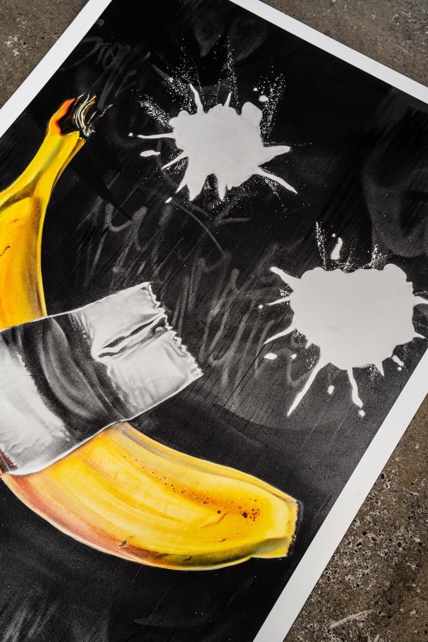 Detailaufnahme eines Graffiti-Art-Prints von Stone Graffiti. Das Motiv zeigt eine realistisch gemalte Banane, die mit silbernem Klebeband auf einem dunklen Hintergrund befestigt zu sein scheint. Darüber sind zwei gesprühte weiße Farbkleckse zu sehen, die dem Werk eine dynamische, experimentelle Ästhetik verleihen. Der hochwertige Druck liegt auf einer rauen Betonoberfläche und hebt sich durch seine satten Farben und feinen Details ab.
