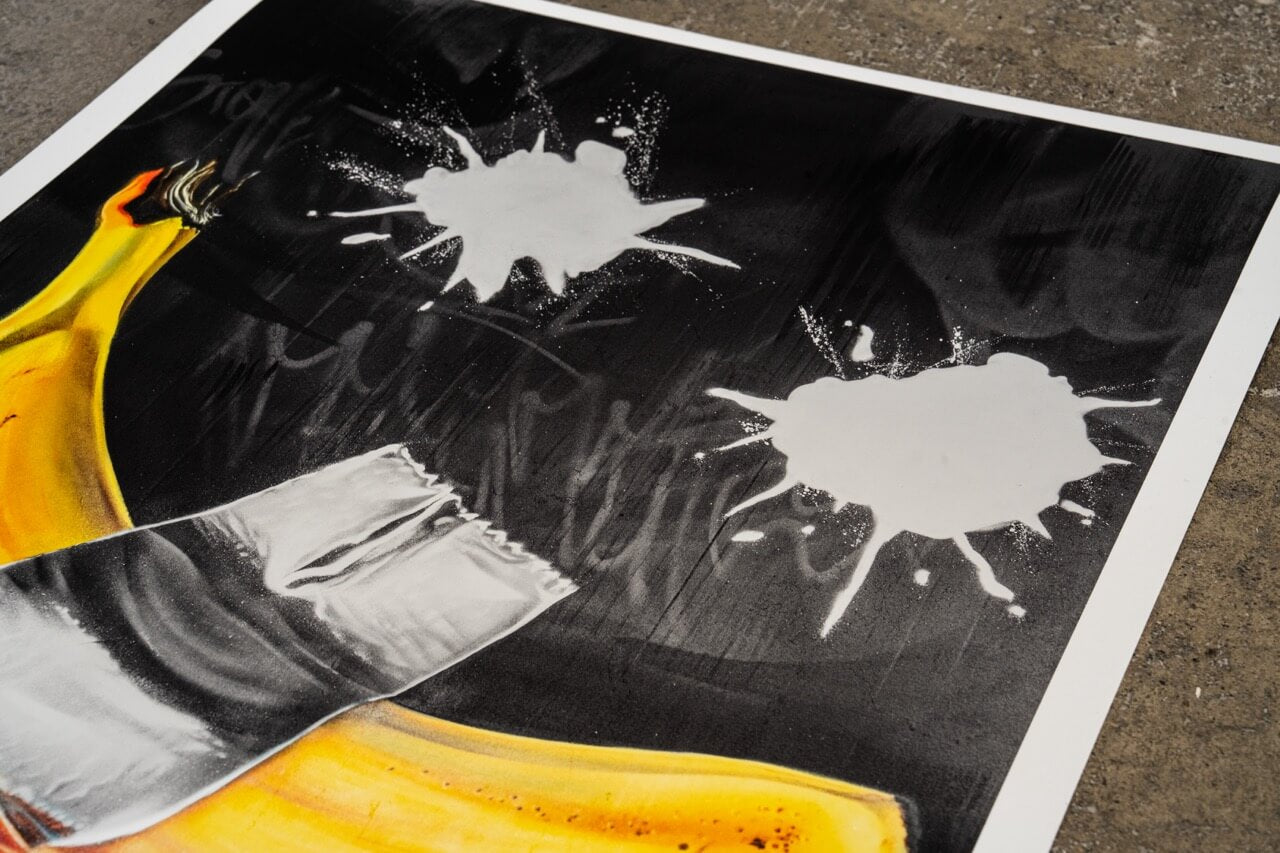 Detailaufnahme eines Graffiti-Kunstdrucks von Stone Graffiti. Das Motiv zeigt eine realistisch gemalte Banane, die mit silbernem Klebeband auf einem dunklen Hintergrund fixiert scheint. Darüber sind zwei große weiße Farbkleckse gesprüht, die dem Werk eine experimentelle und dynamische Note verleihen. Der hochwertige Druck liegt auf einer Betonoberfläche und hebt sich durch seine kräftigen Kontraste und feinen Details ab.