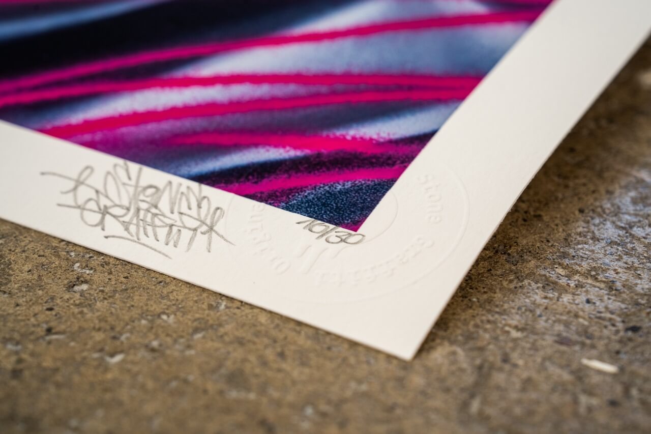 Detailaufnahme einer limitierten Kunstdruck-Edition von Stone Graffiti. Der Ausschnitt zeigt die untere Ecke des hochwertigen Drucks mit Signatur des Künstlers, der Editionsnummer '10/30' und einer geprägten Echtheitsprägung. Die lebendigen Farben, darunter leuchtende Pink- und Blautöne, verlaufen dynamisch über die Leinwand. Der Print liegt auf einer groben Betonfläche, die einen starken Kontrast zur feinen Papierstruktur bildet.