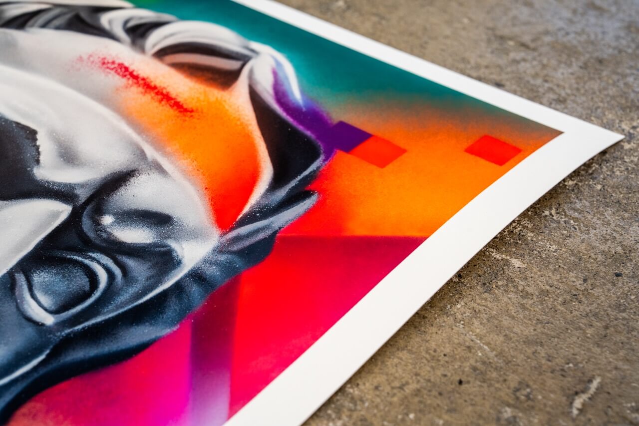 Nahaufnahme eines limitierten Kunstdrucks von Stone Graffiti. Der Bildausschnitt zeigt die farbintensive Ecke des Prints, auf der ein detaillierter, gesprayter Skulpturenkopf in Schwarz-Weiß zu sehen ist. Die umgebenden Farben reichen von leuchtendem Orange über tiefes Rot bis hin zu kräftigem Violett. Digitale Pixel-Elemente in Lila und Rot verstärken den modernen Stil des Kunstwerks. Der Druck liegt auf einer rauen Betonoberfläche, die im Kontrast zur glatten Papierstruktur steht.