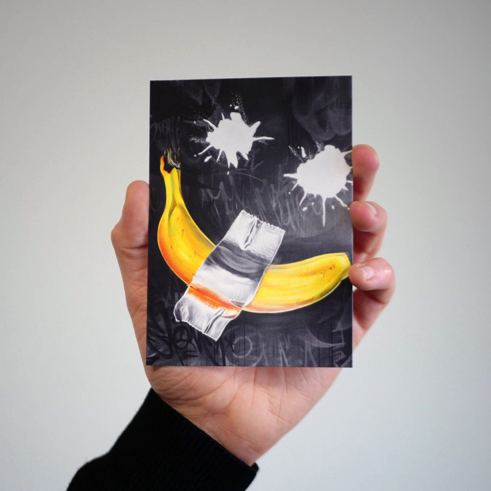 Eine Hand hält eine Postkarte mit einem urbanen Graffiti-Motiv von Stone Graffiti, das eine mit silbernem Klebeband fixierte Banane auf schwarzem Hintergrund zeigt. Die Detailgenauigkeit der gedruckten Kunst und die kräftigen Farben kommen in der Nahaufnahme besonders gut zur Geltung.